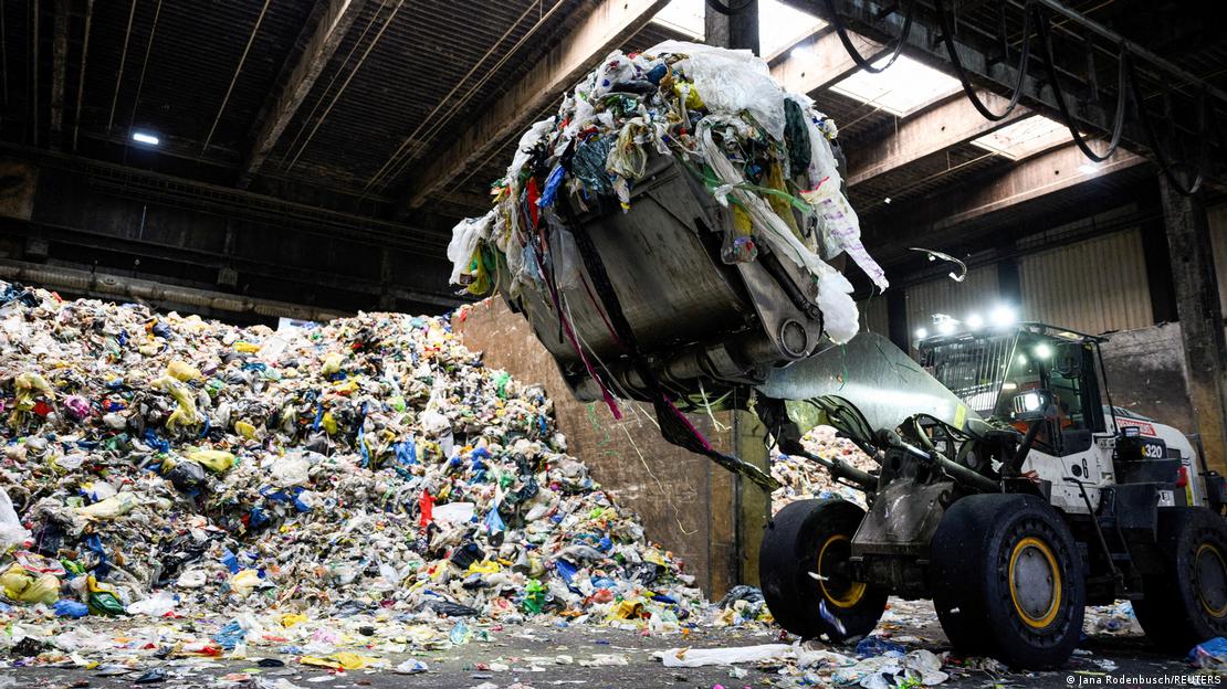 Bir atık tesisinde, iş makinesi kepçe ile havaya kaldırılan çöp yığını, arka planda büyük bir atık yığını